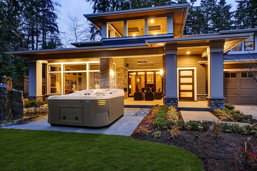 A hot tub beside a large house at dusk with warm light in the windows and a clipped lawn