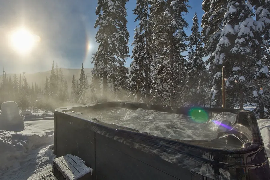 Steam lifting off a hot tub surrounded by deep snow, low sun behind snow-covered pines