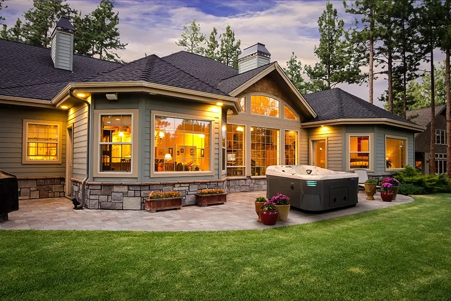 A hot tub on a lit stone patio behind a Front Range home at dusk, with pines behind the roofline